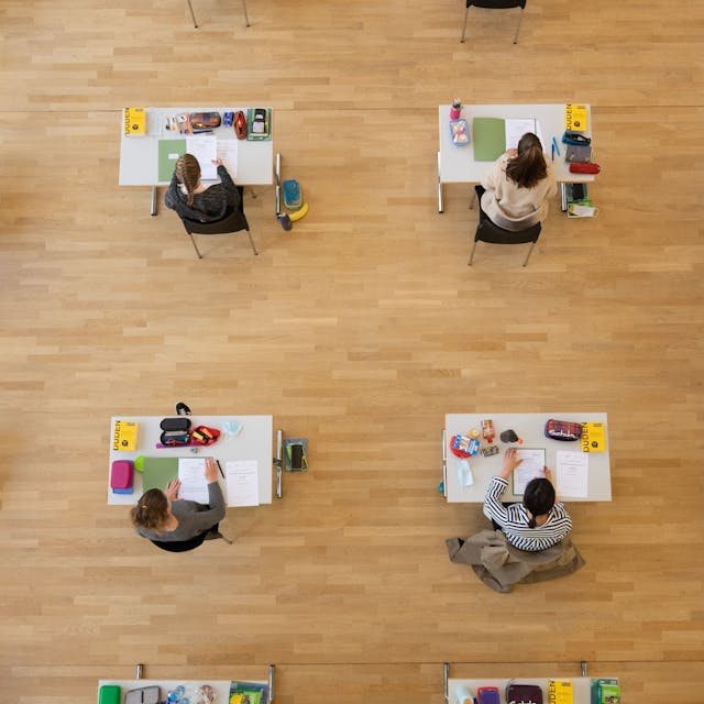 Schüler sind während der Abiturprüfung aus der Vogelperspektive zu sehen. Sie sitzen an weißen Tischen, auf denen je ein Duden liegt, Schreibzeug und die Aufgabenmappe.