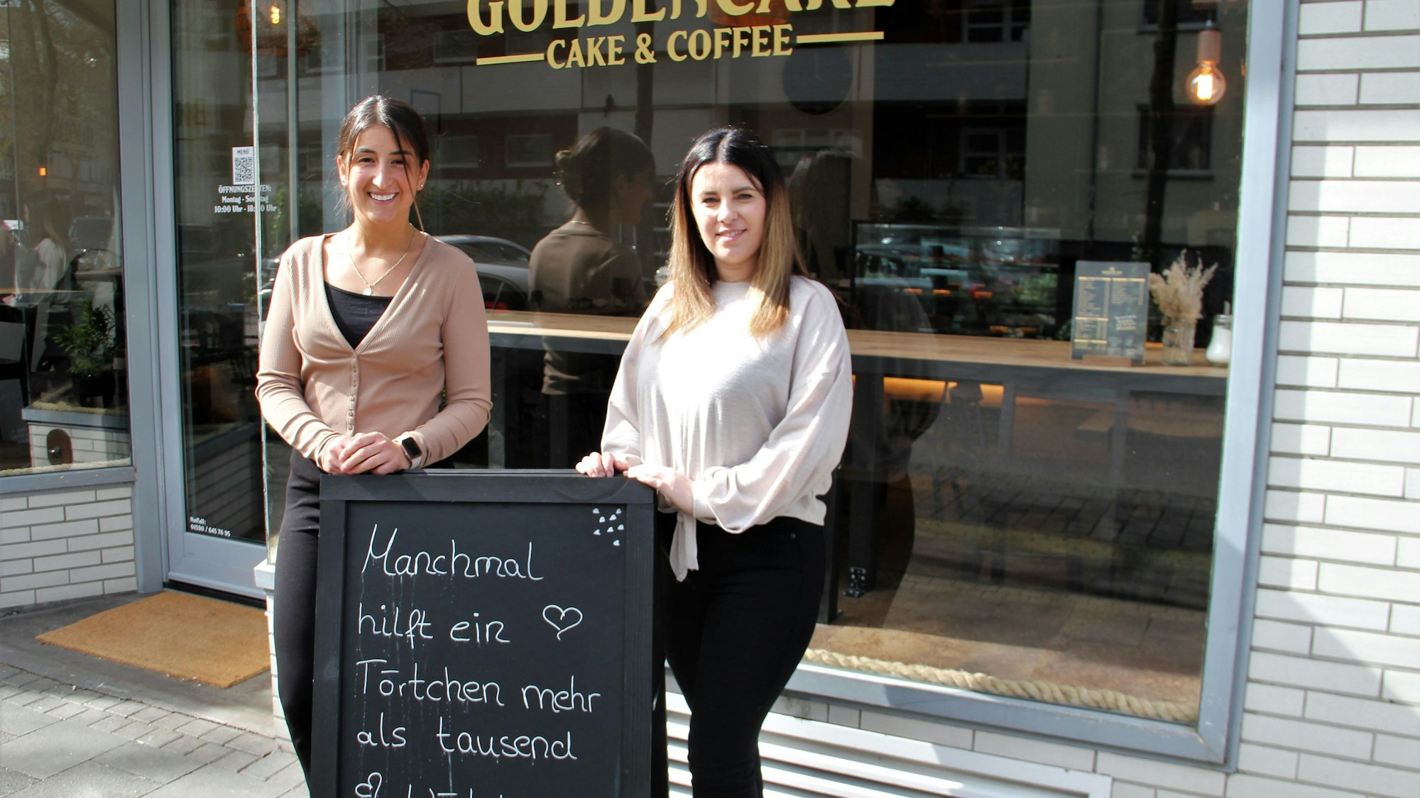 Zwei Frauen stehen vor der Glasfront eines Cafés. Sie stehen hinter einer Tafel, auf der steht Manchmal hilft ein Törtchen mehr als tausend Wörtchen.