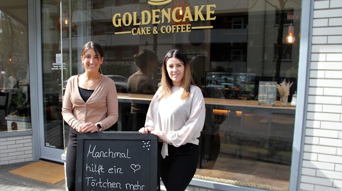 Zwei Frauen stehen vor der Glasfront eines Cafés. Sie stehen hinter einer Tafel, auf der steht Manchmal hilft ein Törtchen mehr als tausend Wörtchen.&nbsp;