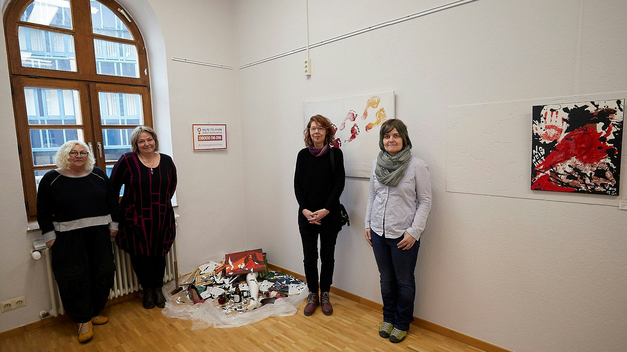 Bilder in der Galerie Eifel Kunst werden gezeigt sowie wie vier Frauen neben einem Kunstwerk über hausliche Gewalt.
