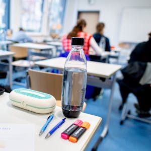 Ein Klassenraum mit mehreren Schülerinnen und Schülern, im Vordergrund ein Schreibtisch mit einem Bogen, auf dem „Abitur 2023“ steht.