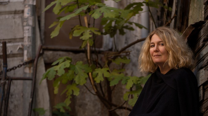 Sabine Schiffner sitzt vor einer grauen, von einer Pflanze berankten, Außenwand. Sie trägt ein schwarzes Oberteil und blickt in die Kamera