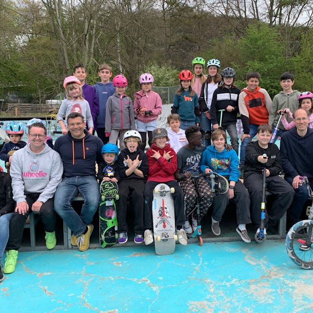 Eine Gruppe von Kindern, Jugendlichen und Erwachsenen haben sich mit Rollern, Skateboards und Fahrrädern zum Gruppenbild postiert. Viele Kinder tragen Fahrradhelme.