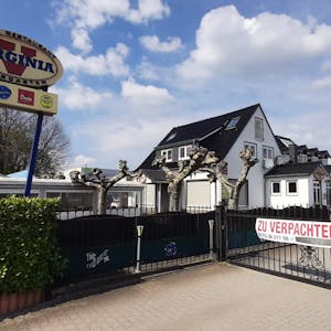 Das Virginia American Bar Restaurant in Monheim am Rhein befindet sich mittig hinter einem Zaun. Links findet sich ein Werbeschild mit dem Logo des Restaurants, rechts der Schotterparkplatz ohne Autos.