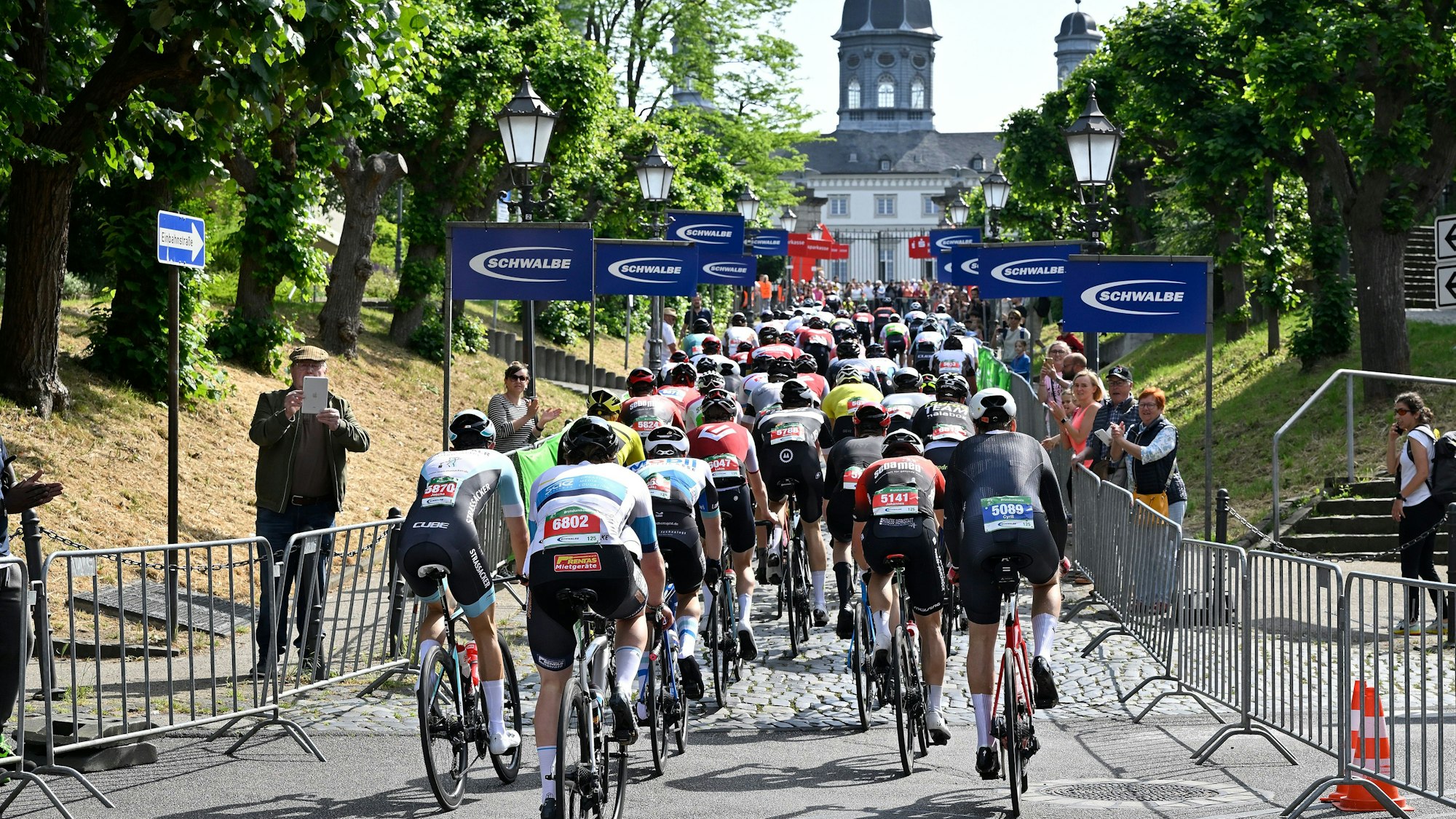 Radrennfahrer fahren die Kopfsteinpflasterstraße zum Bensberger Schloss hinauf. Neben der Fahrbahn stehen Zuschauer hinter Drängelgittern.