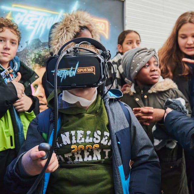 Ein Junge hat eine VR-Brille auf, auf der „Beethoven Opus 360 “steht. Um ihn herum haben sich Mitschülerinnen und Mitschüler versammelt.