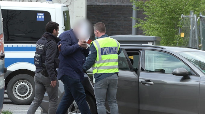 Zwei Männer, die Westen mit der Aufschrift „Polizei“ tragen, führen einen Mann in Handschellen zu einem Auto.&nbsp;