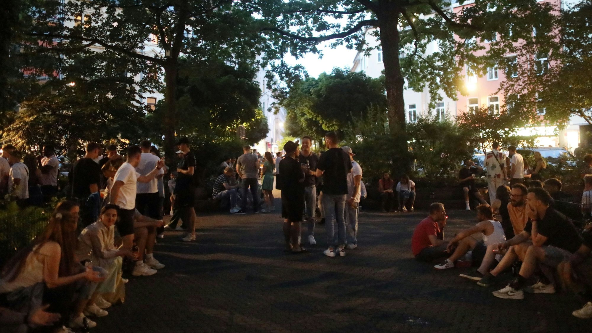 Menschen stehen und sitzen auf einem mit Bäumen umgebenen Platz