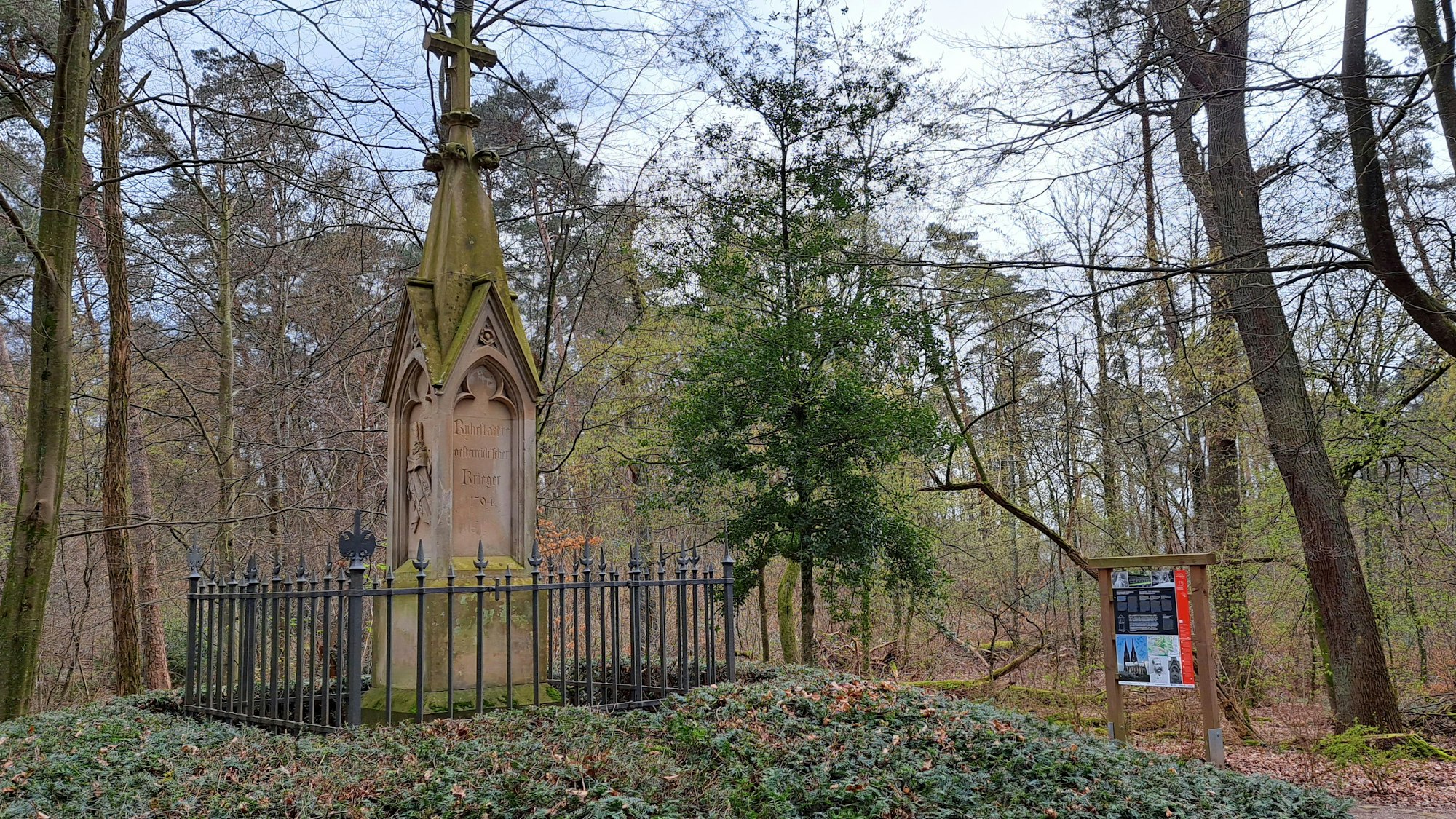 Ein Denkmal steht umzäunt in einem Wald, daneben eine bebilderte Informationstafel.