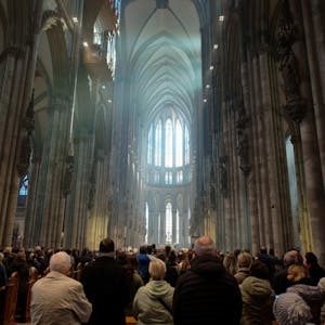 Blick in den Kölner Dom während einer Messe. Die Menschenmenge ist von hinten zu sehen, der Blick geht nach oben zu den gotischen Fenstern im Chor, die von Tageslicht erhellt sind.