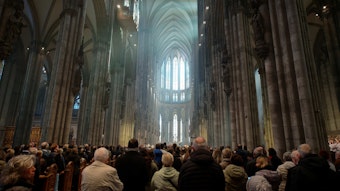 Blick in den Kölner Dom während einer Messe. Die Menschenmenge ist von hinten zu sehen, der Blick geht nach oben zu den gotischen Fenstern im Chor, die von Tageslicht erhellt sind.