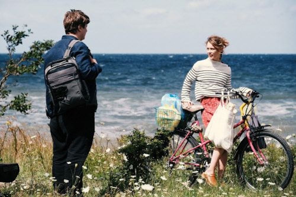 Eine junge Frau mit Fahrrad spricht mit einem jungen Mann, im Hintergrund sieht man das Meer.