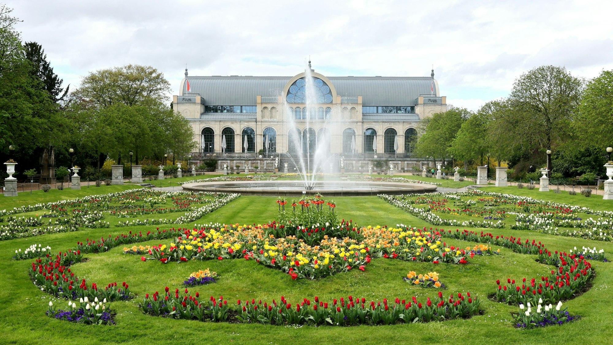 Der Glaspalast in der Kölner Flora mit dem Springbrunnen und Blumenbeeten im Bildvordergrund