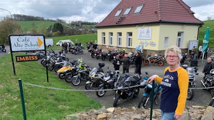 Vor acht Jahren hat Annette Büth die Verantwortung für den Bikertreff übernommen. Eine Frau mit blonden kurzen Haaren steht vor dem Café Alte Schule, auf dem Parkplatz stehen viele Motorräder.