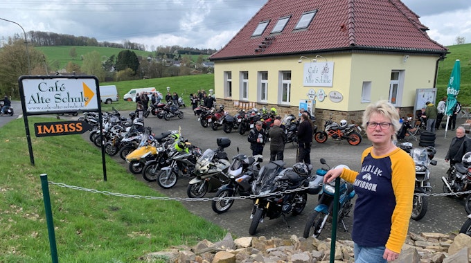 Vor acht Jahren hat Annette Büth die Verantwortung für den Bikertreff übernommen. Eine Frau mit blonden kurzen Haaren steht vor dem Café Alte Schule, auf dem Parkplatz stehen viele Motorräder.