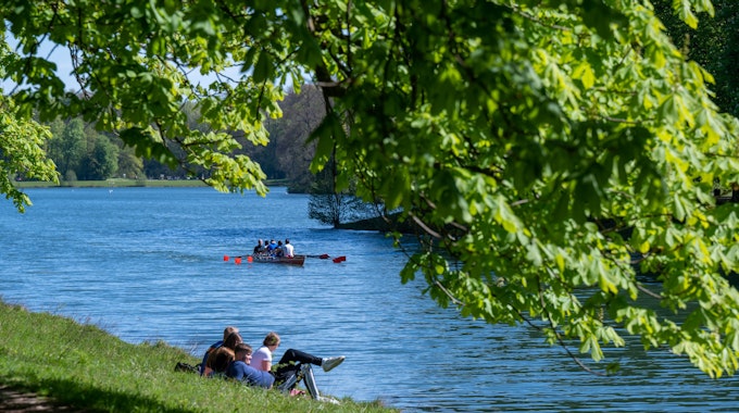 Menschen sitzen am Ufer eines Kanals, die Bäume sind grün, das Wasser blau, ein Ruderboot ist in der Ferne zu sehen.