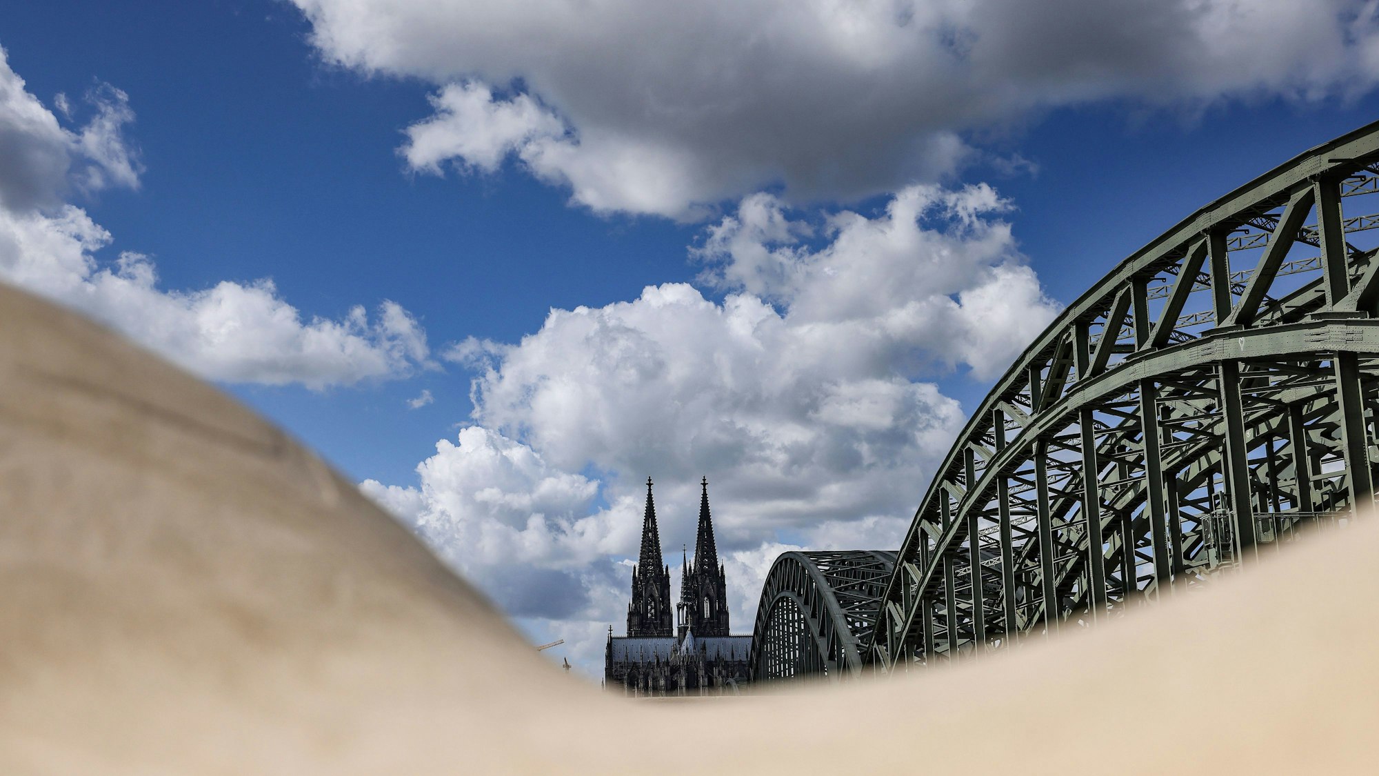 Köln: Wolken ziehen über Dom und Hohenzollernbrücke hinweg, die hinter rundlich geformten Sitzelementen zu sehen sind. Anfang Mai könnte es Sonne im Rheinland geben.