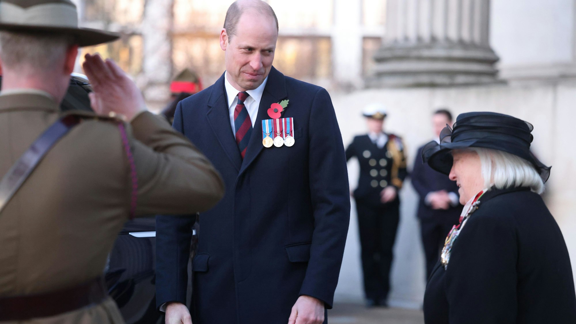 Prinz William, Prinz von Wales, nimmt an der Morgenandacht zum Anzac Day 2023 teil. Er unterhält sich mit einer Teilnehmerin. Vor ihm steht ein salutierender Soldat.