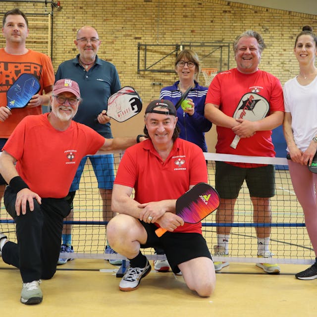 Fünf Männer und zwei Frauen unterschiedlichen Alters in Sportkleidung in einer Turnhalle vor einem Netz mit Schlägern in der Hand