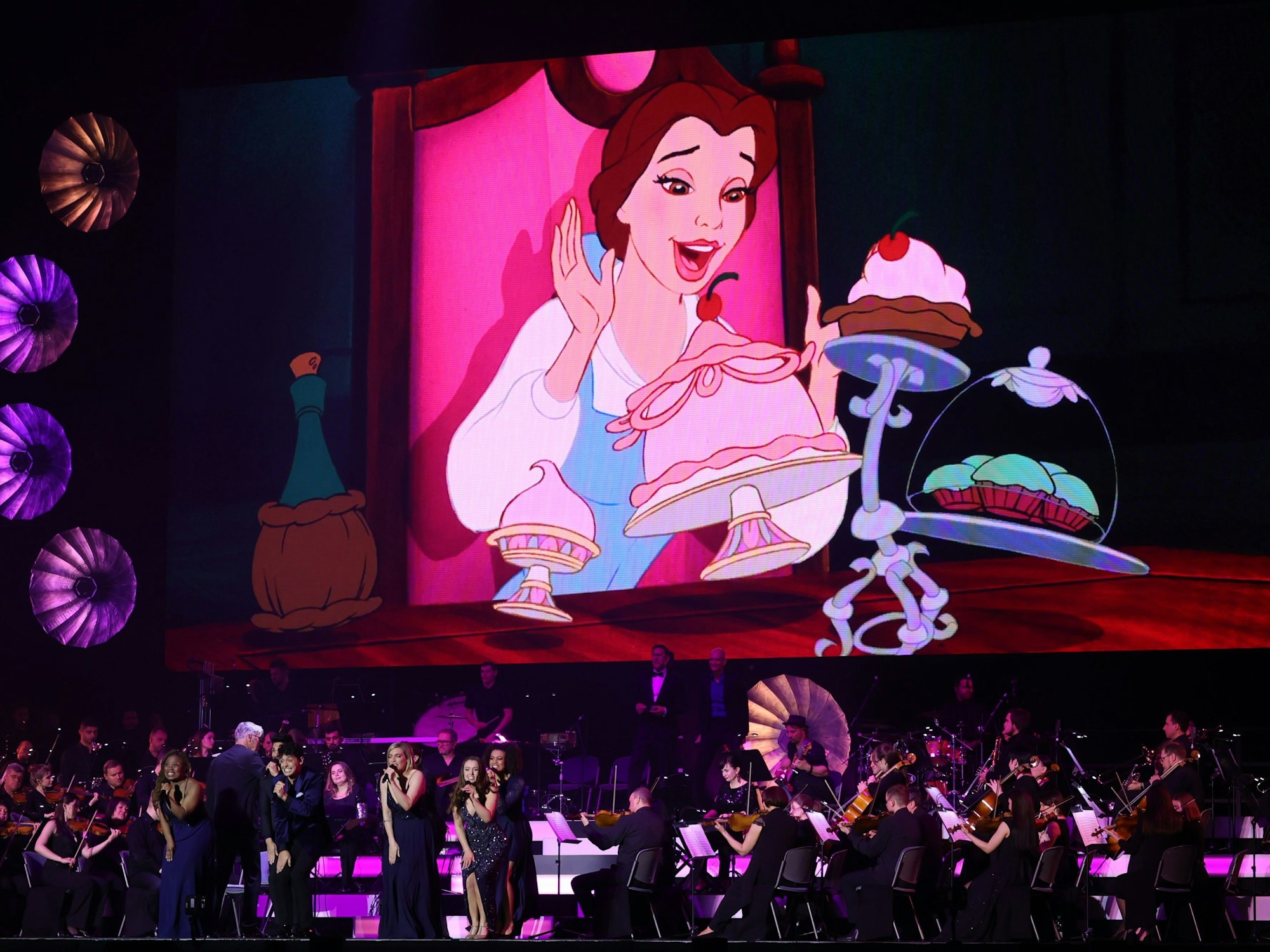 Das Orchester spielt auf der Bühne während man im Hintergrund eine Szene aus „Die Schöne und das Biest“ sieht.