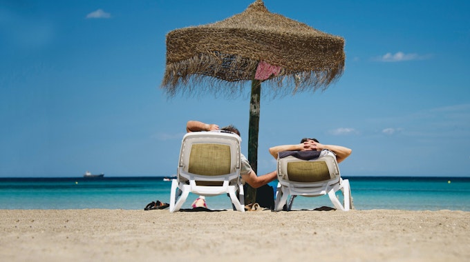 Zwei Urlauber auf Liegen unter einem Sonnenschirm an der&nbsp;Plaja de Muro auf Mallorca.