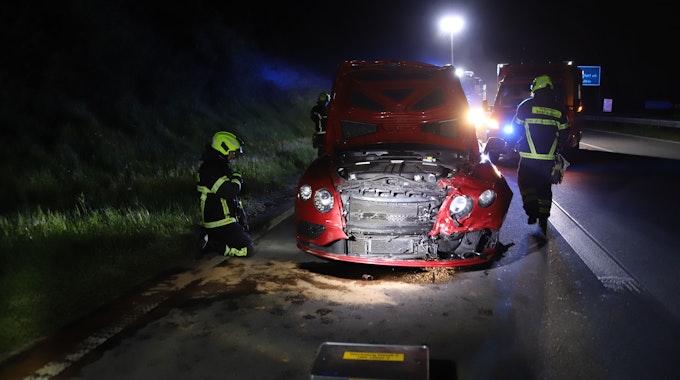 Feuerwehrkräfte begutachten nachts ein rotes Auto nach einem Unfall. Die Front ist von einer Kollision beschädigt.