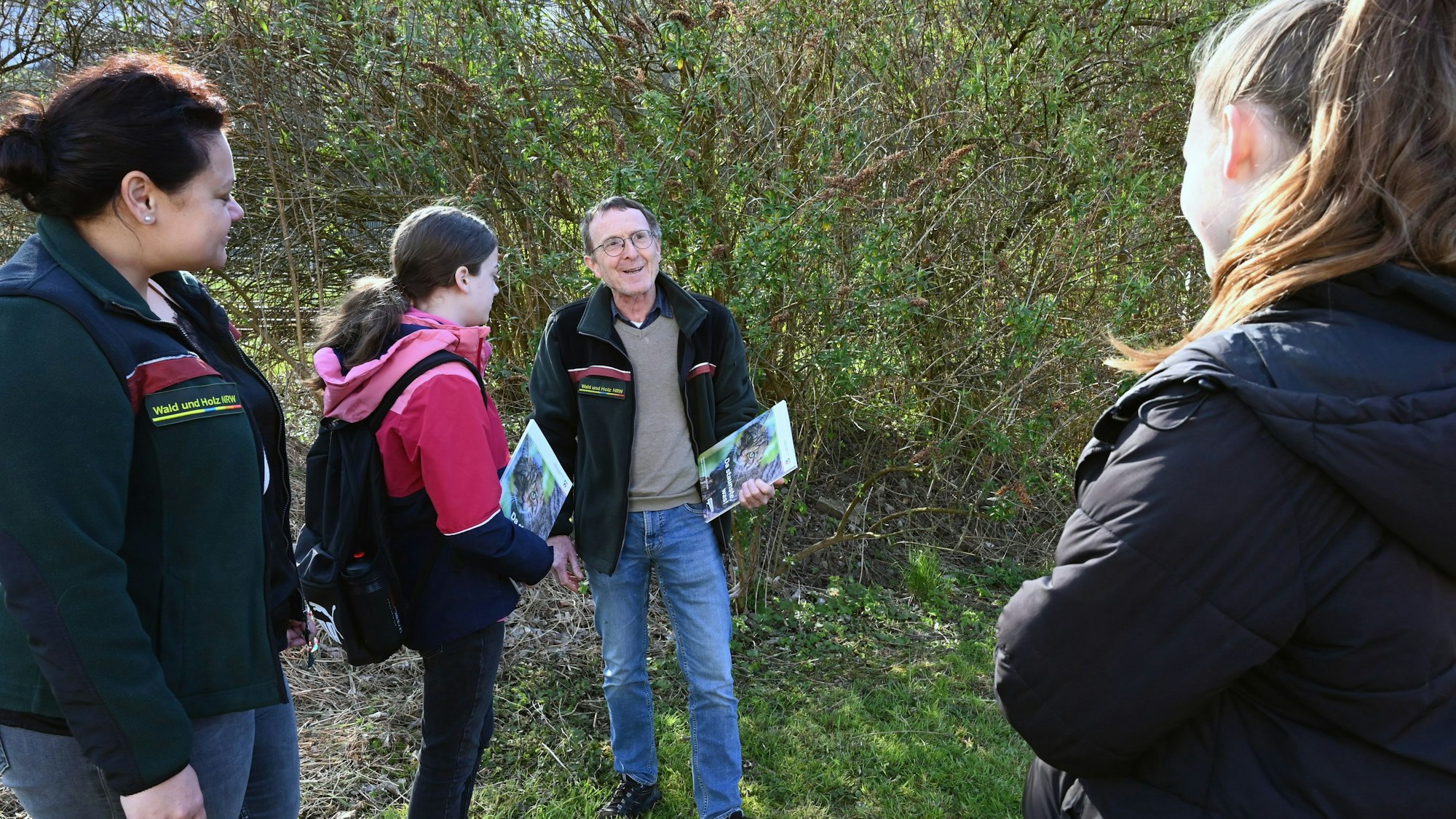 Ein Mann steht mit mehreren Schülerinnen im Wald. Er hat eine Informationsmappe in der Hand und spricht zu den jungen Frauen.