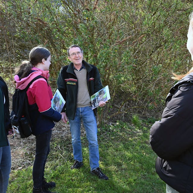 Ein Mann steht mit mehreren Schülerinnen im Wald. Er hat eine Informationsmappe in der Hand und spricht zu den jungen Frauen.