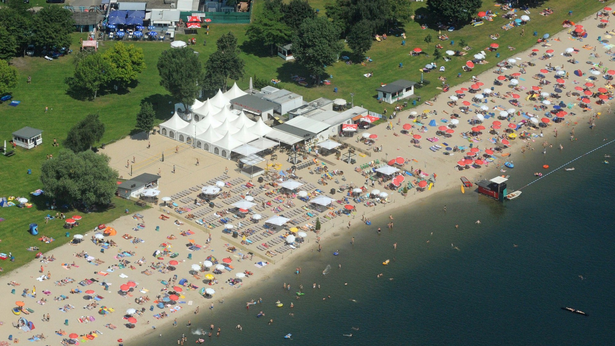 Am Strandbach liegen viele Menschen auf Handtüchern. Weiße Zelte stehen am Strand.