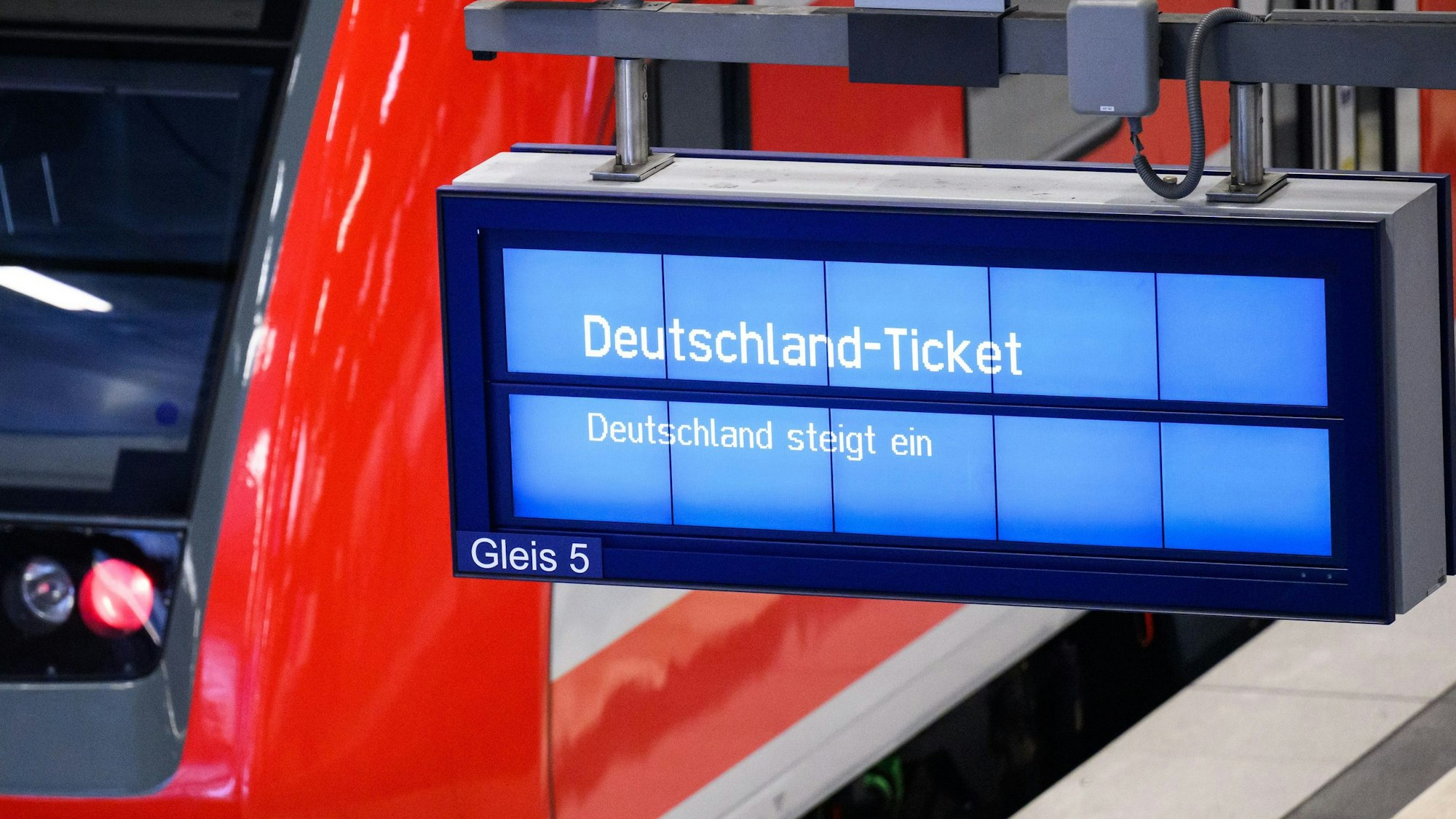 Auf einer Anzeige an einem Bahnhofsgleis steht groß „Deutschland-Ticket“. Im Hintergrund ist ein rot-weißer Regionalzug der Deutschen Bahn zu sehen.