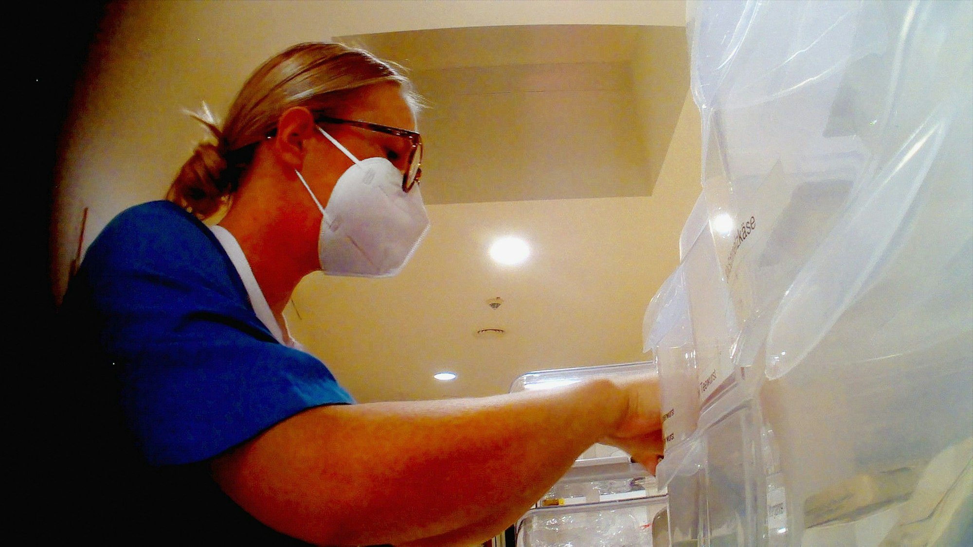 Eine junge Frau mit blondem Zopf und Brille in blauem medizinischen Kaftan und FFP2-Maske sortiert medizinisches Material in Plastikboxen.