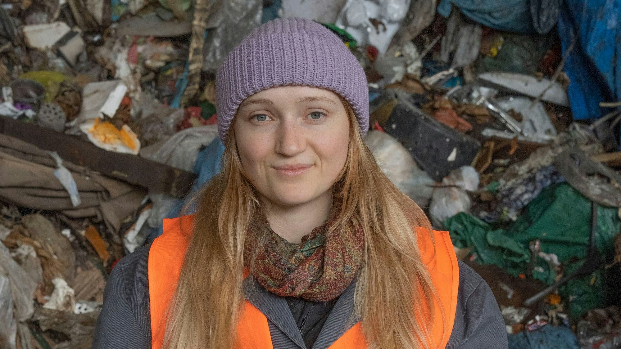 Eine Studentin der TH Köln steht vor einem Berg aus Müll. Sie trägt eine lila Mütze und eine orange Warnweste.