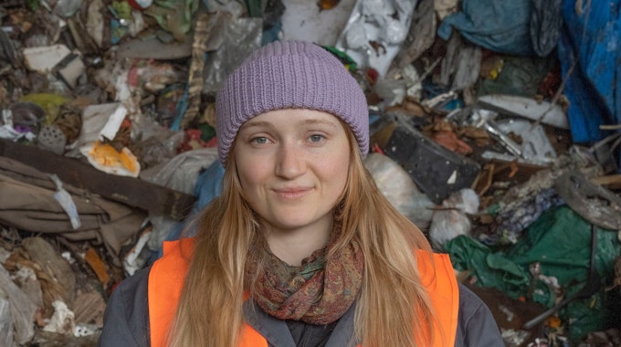 Eine Studentin der TH Köln steht vor einem Berg aus Müll. Sie trägt eine lila Mütze und eine orange Warnweste.&nbsp;