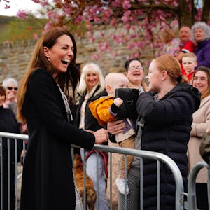 Kate Middleton lacht, während ein Baby mit ihrer Handtasche spielt. Seine Mutter hält das Baby auf dem Arm.