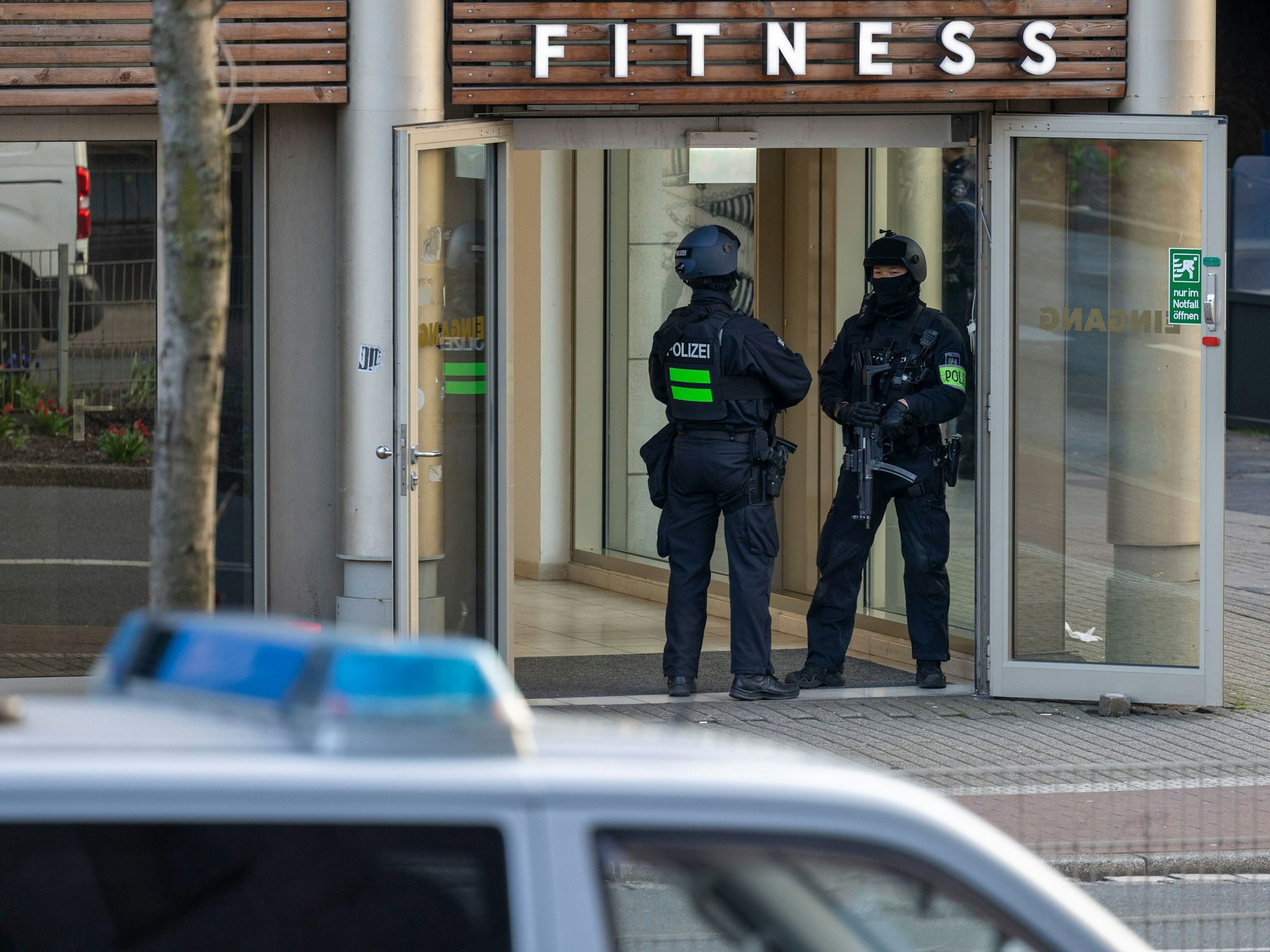 Das Foto vom 18. April 2023 zeigt zwei Polizisten, die im Eingang eines Fitnessstudios stehen. Im Vordergrund befindet sich ein Einsatzwagen der Polizei.