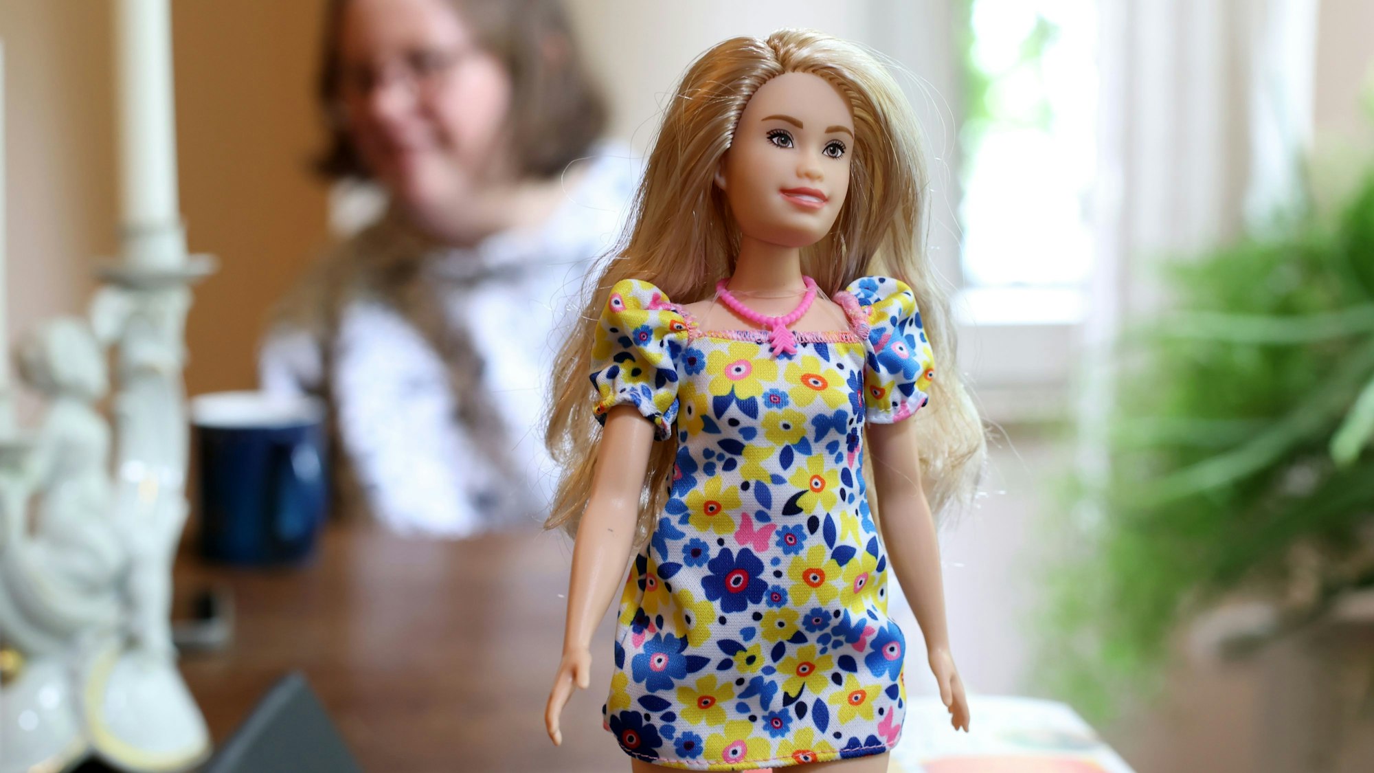 Das Bild zeigt die erste Barbie mit Down-Syndrom. Sie hat blonde, lange Haate, trägt ein Kleid mit blauen und gelben Blumen und eine rosafarbene Kette.