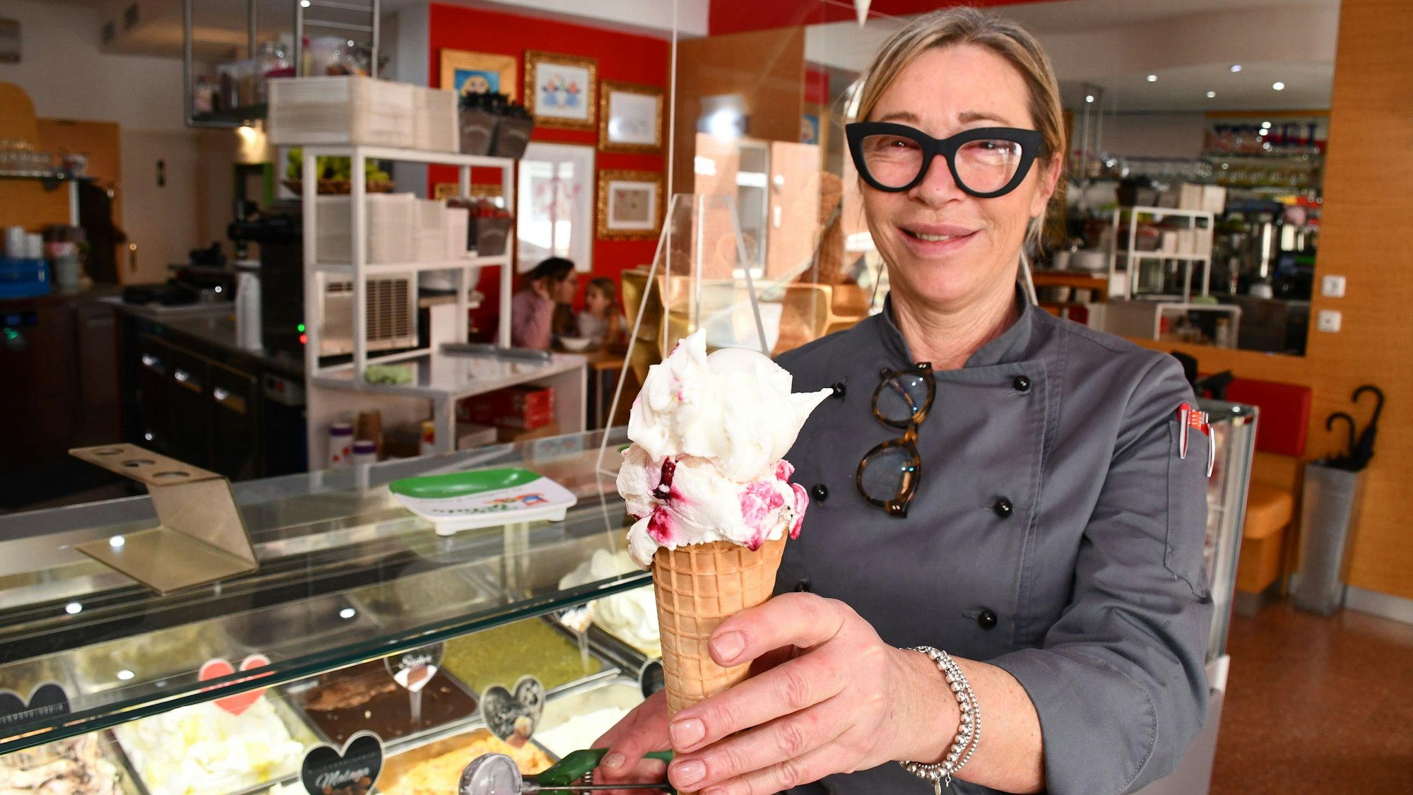 Eine Frau mit Brille hält in einer Eisdiele lächelnd ein Waffel-Eis in die Kamera.