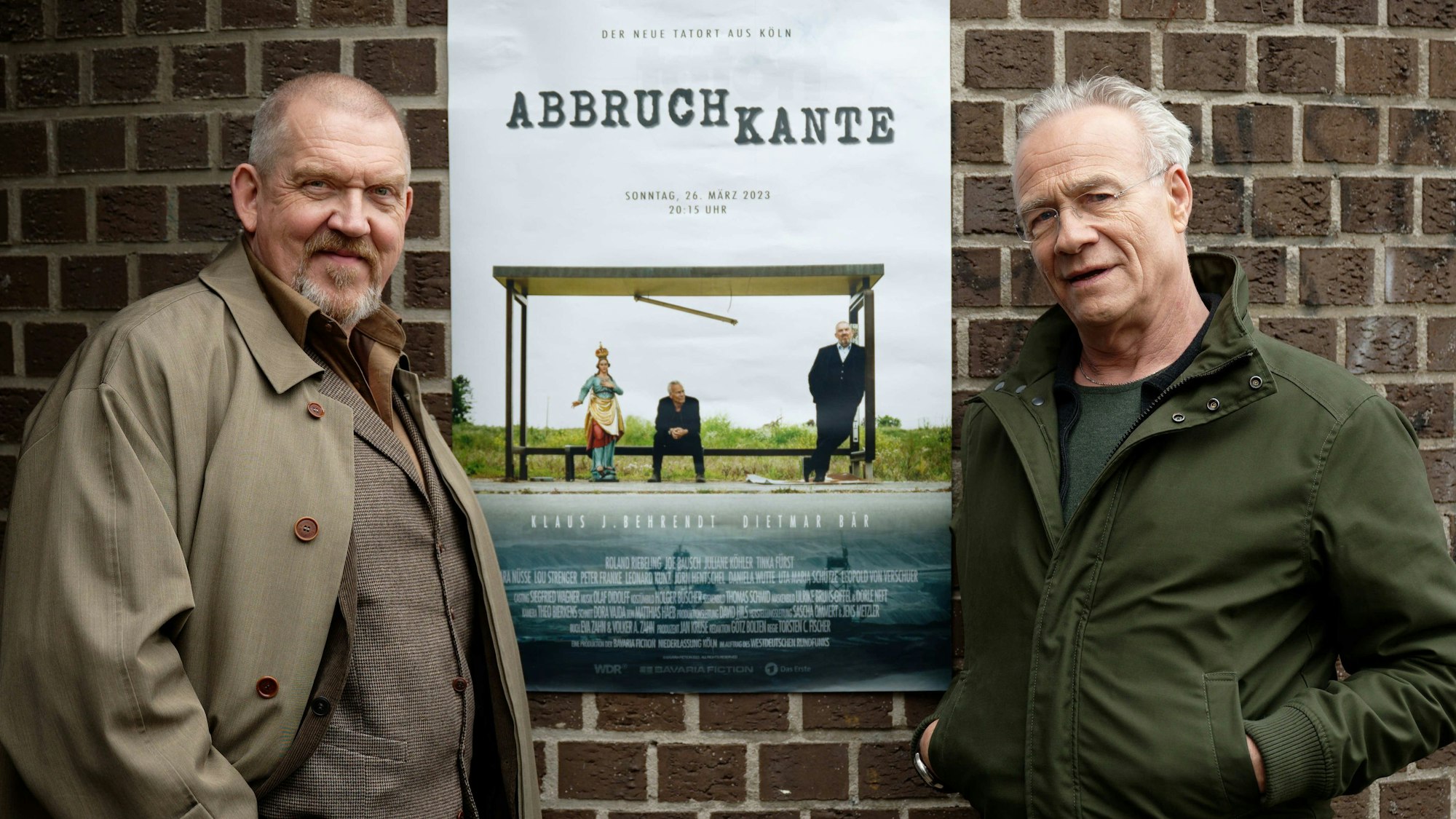 Die Schauspieler Klaus J. Behrendt und Dietmar Bär stehen bei einem Pressetermin bei Dreharbeiten vor einem Plakat.