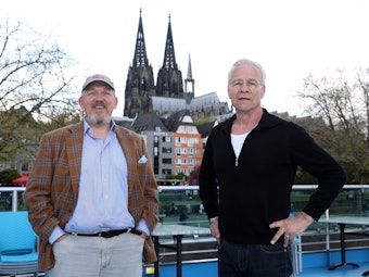 Dietmar Bär und Klaus J. Behrendt stehen auf einem Schiff am Rheinufer.