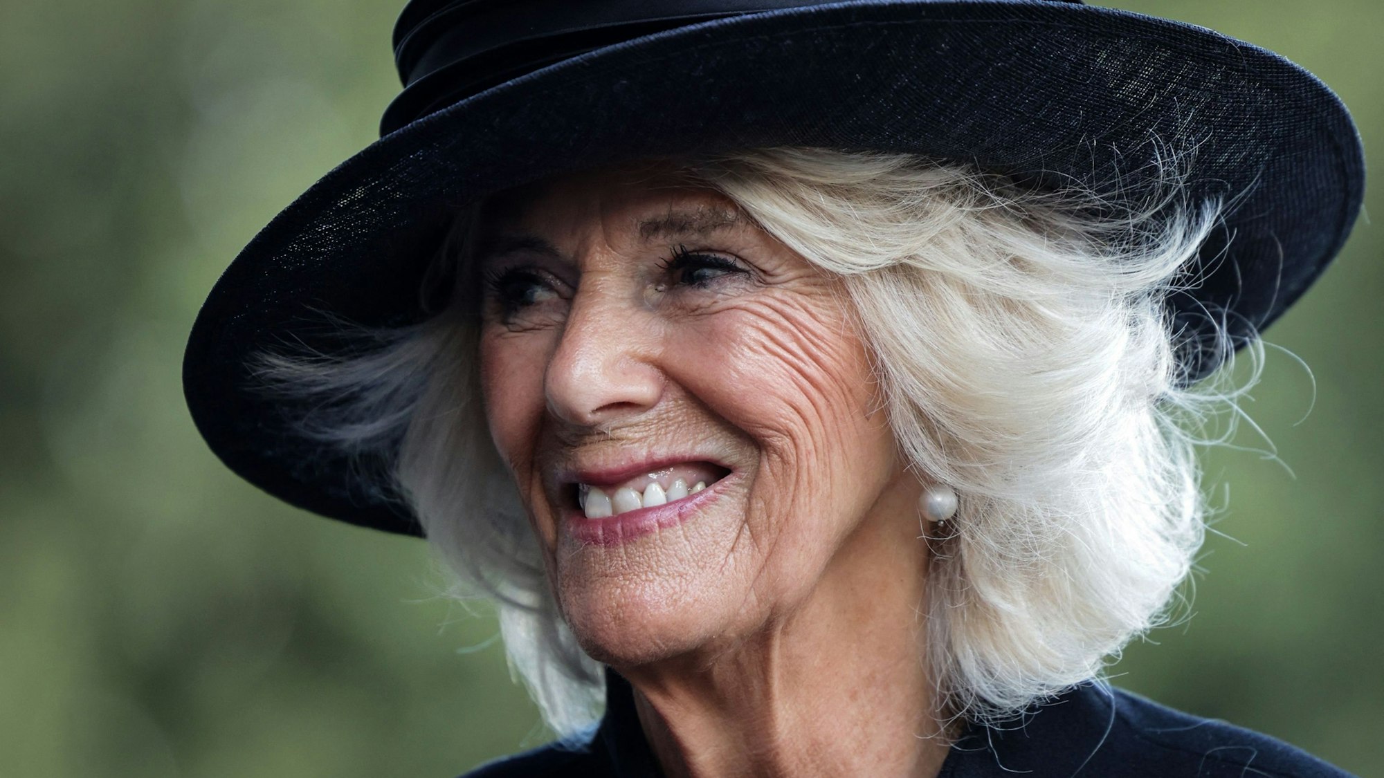 Königin Camilla lächelt. Sie trägt ein schwarzes Kleid und einen schwarzen Hut.