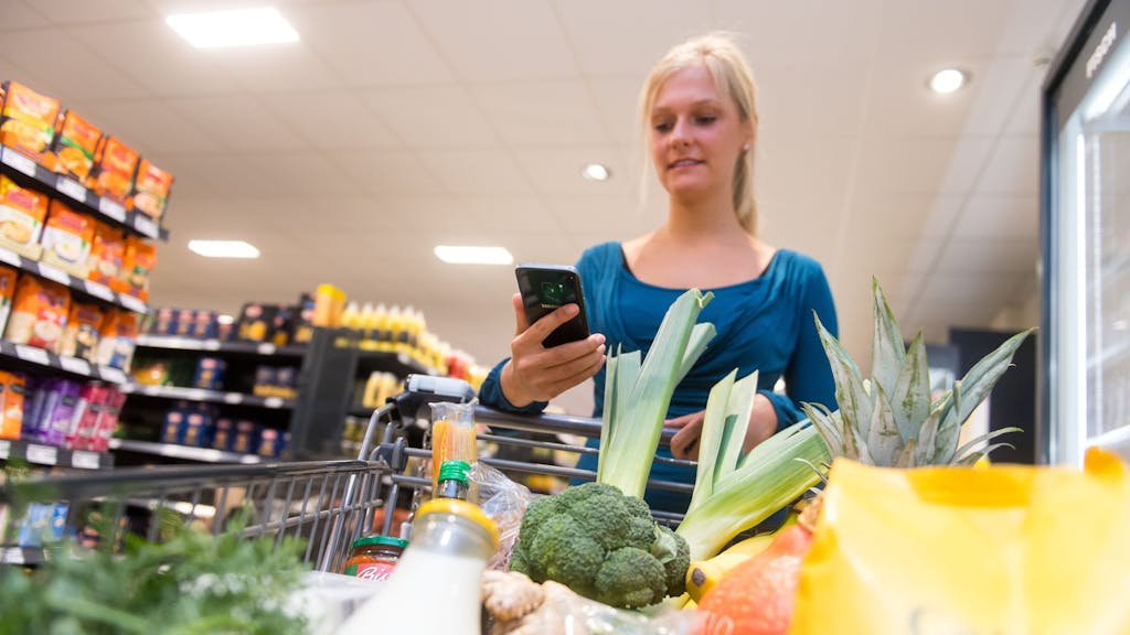 Eine Frau kauft in einem Supermarkt mit einem Einkaufswagen ein. In ihrer Hand hält sie ein Smartphone.&nbsp;