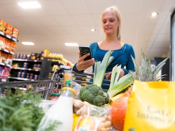 Eine Frau kauft in einem Supermarkt mit einem Einkaufswagen ein. In ihrer Hand hält sie ein Smartphone.