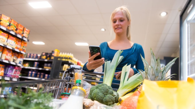 Eine Frau kauft in einem Supermarkt mit einem Einkaufswagen ein. In ihrer Hand hält sie ein Smartphone.&nbsp;