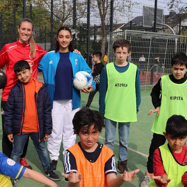 Ex-FC Profispielerin Nina Windmüller steht vor einem Fußballtor umgeben von Kindern in bunten Trikots.