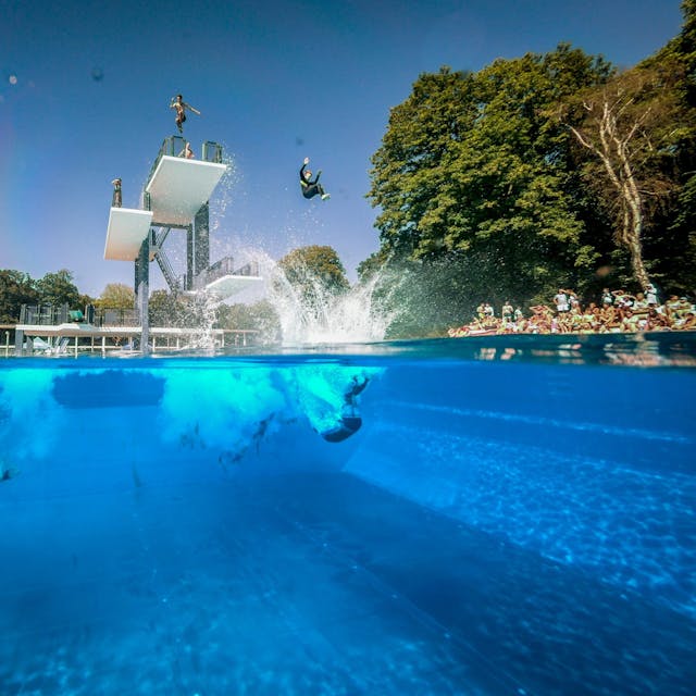 Menschen springen vom Sprungturm im Freibad Stadionbad in Köln ins Wasser.