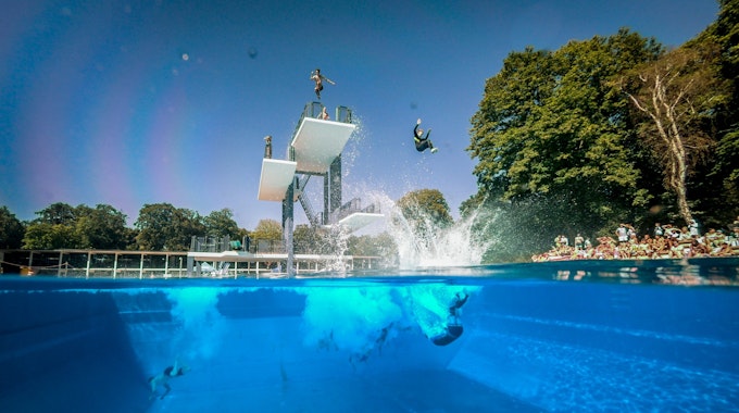 Menschen springen vom Sprungturm im Freibad Stadionbad in Köln ins Wasser.
