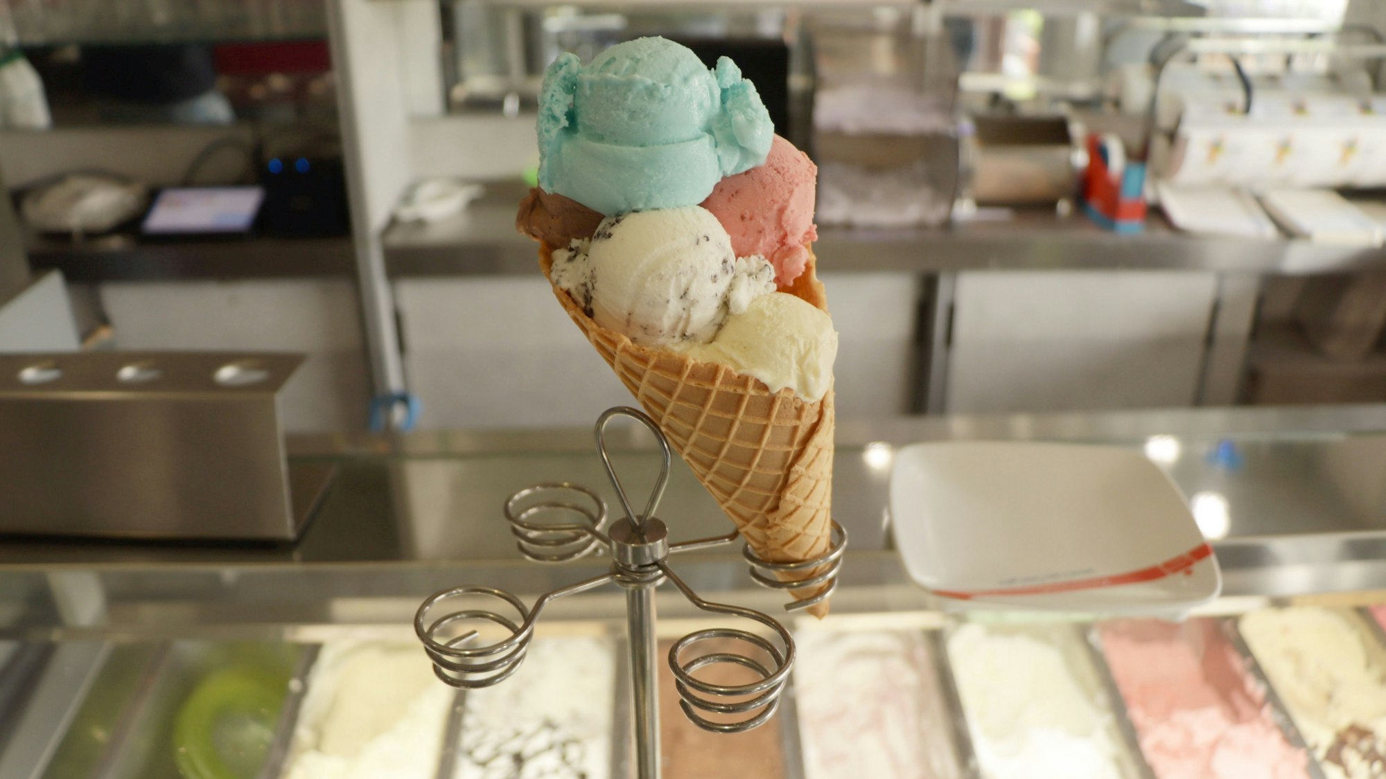 Eine große Eiswaffel mit mehreren Eiskugeln ist mittig im Bild platziert. Es befindet sich auf einem Eiswaffelständer an der Theke eines Eiscafés.