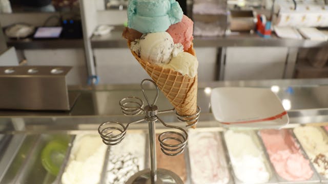 Eine große Eiswaffel mit mehreren Eiskugeln ist mittig im Bild platziert. Es befindet sich auf einem Eiswaffelständer an der Theke eines Eiscafés.