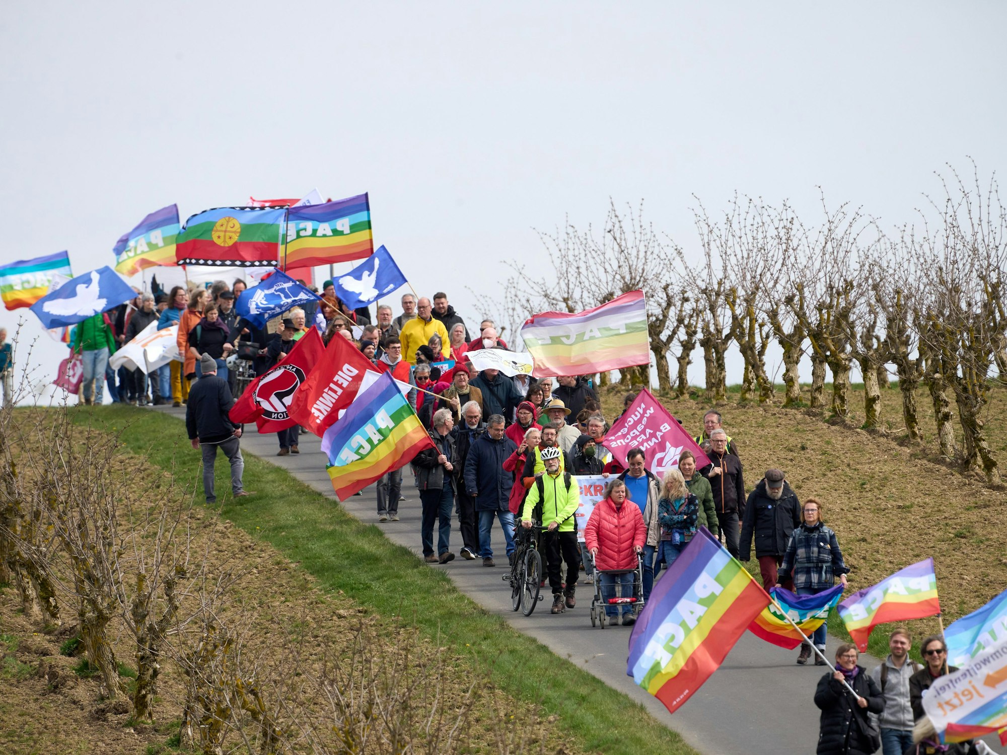 Teilnehmer des Ostermarsches laufen am Zaun des Bundeswehr-Fliegerhorsts mit bunten Flaggen vorbei.