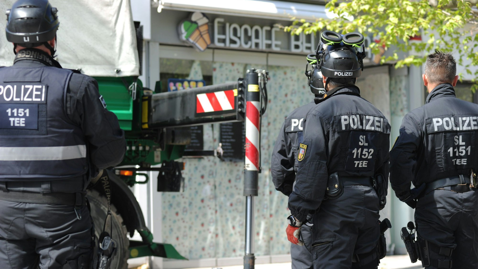 Polizisten sichern im Rahmen einer Razzia Beweismittel. Zu sehen sind Einsatzkräfte, auf deren Rücken "Polizei" zu lesen ist. Sei stehen vor einem Eiscafé.